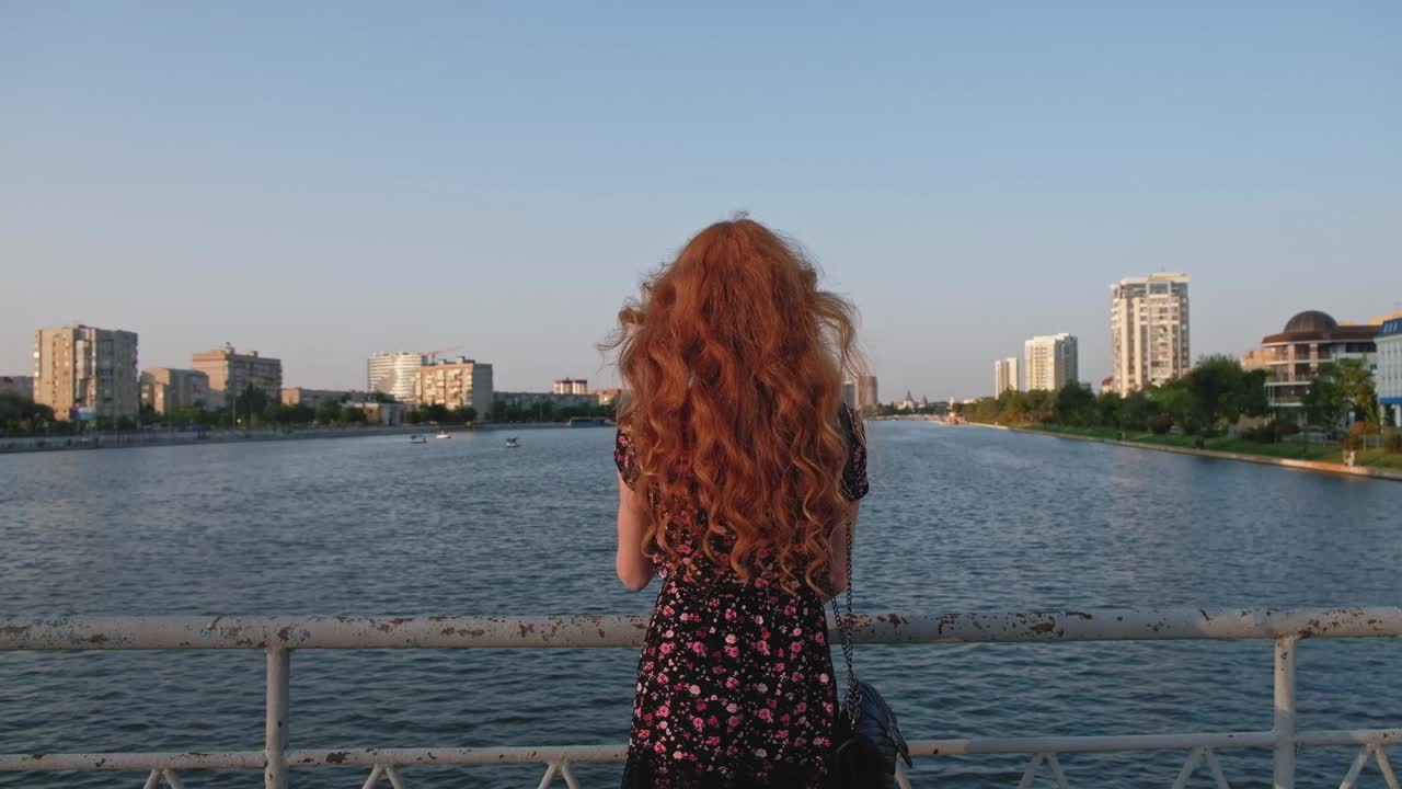 红发女孩穿着夏装从阿斯特拉罕河上的桥看俄罗斯的后景视频下载