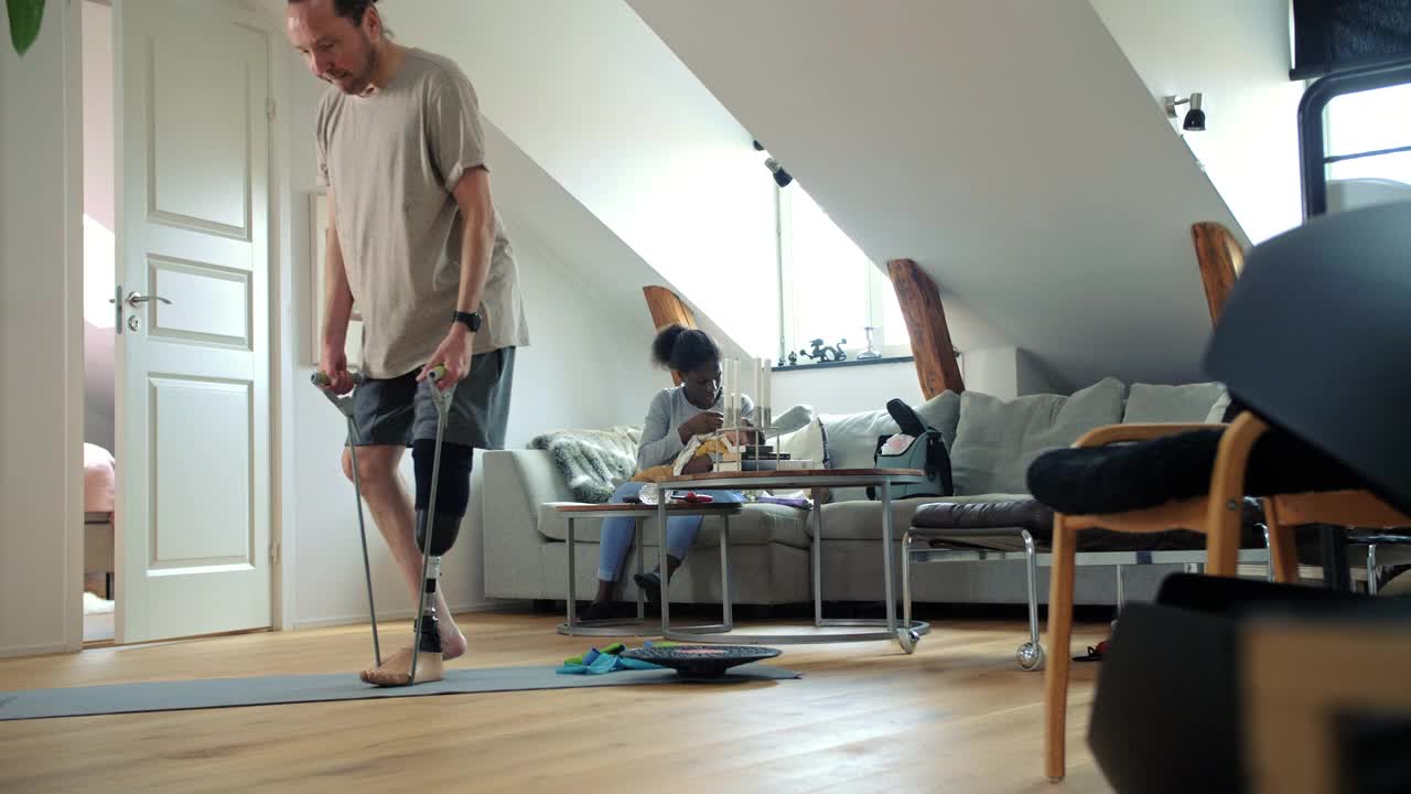 一个带假肢的男人用阻力带运动，而家人坐在家里的沙发上视频下载