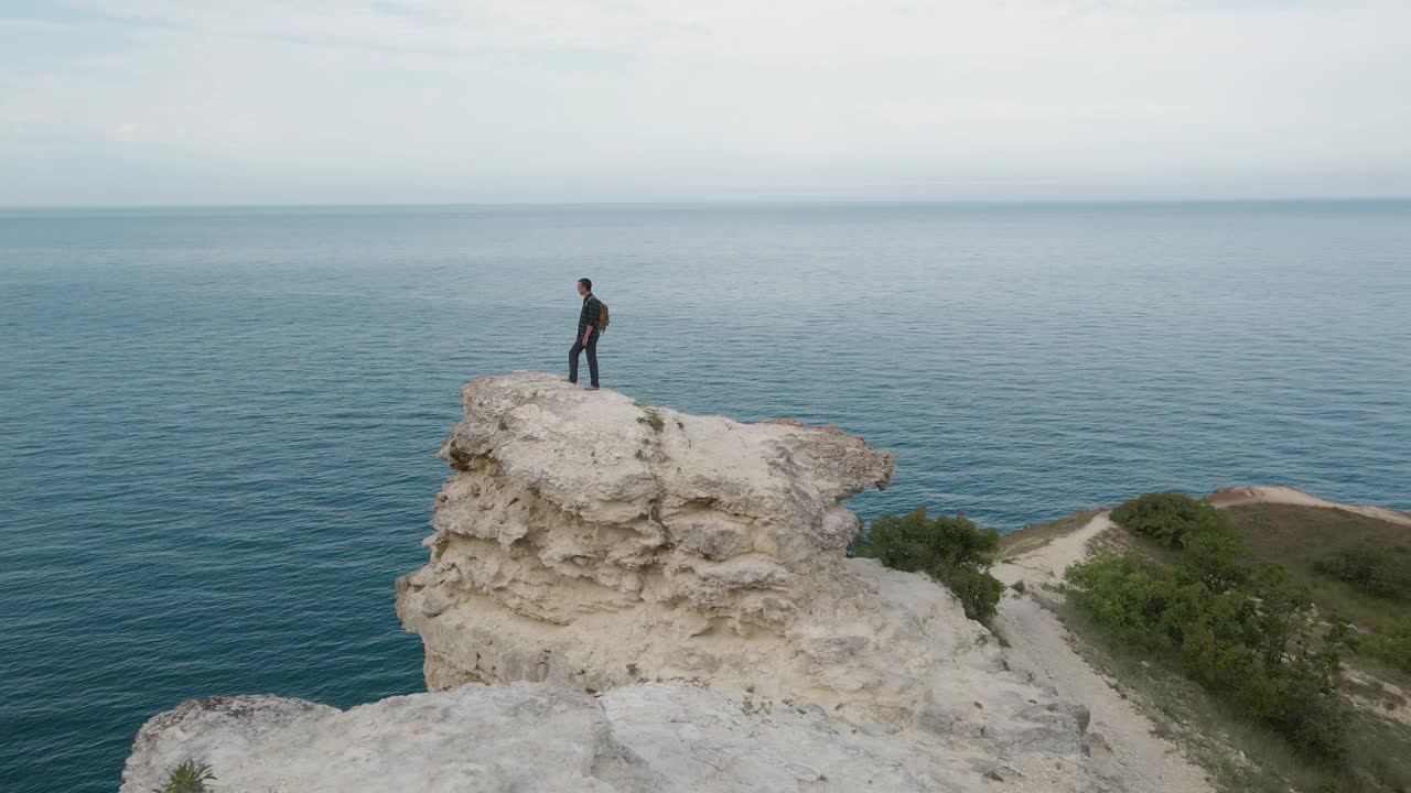 在户外休闲、运动、旅游、娱乐活动中，背包徒步者站在靠近蓝色海洋、天空地平线的岩石山上的无人机旋转视图视频下载
