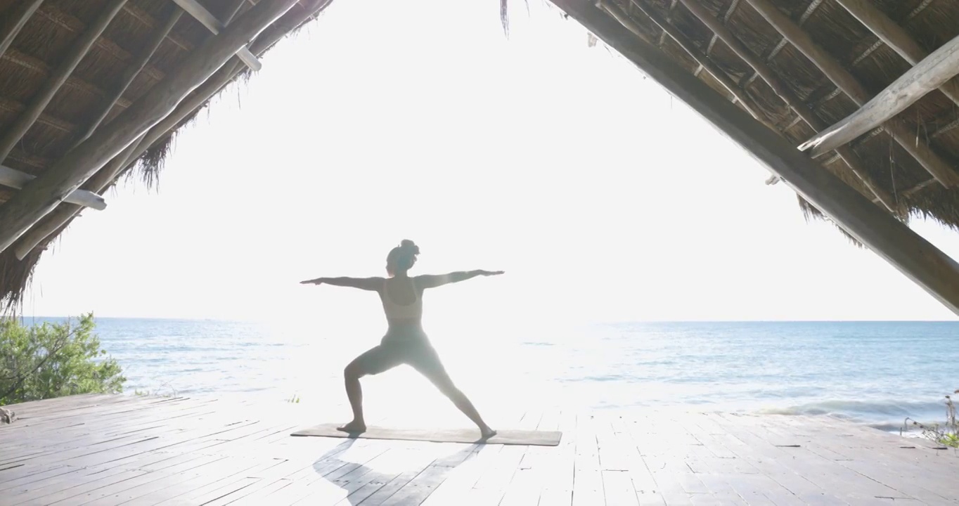 在热带度假胜地的海滨凉亭练习瑜伽时，在勇士姿势的妇女宽镜头视频素材