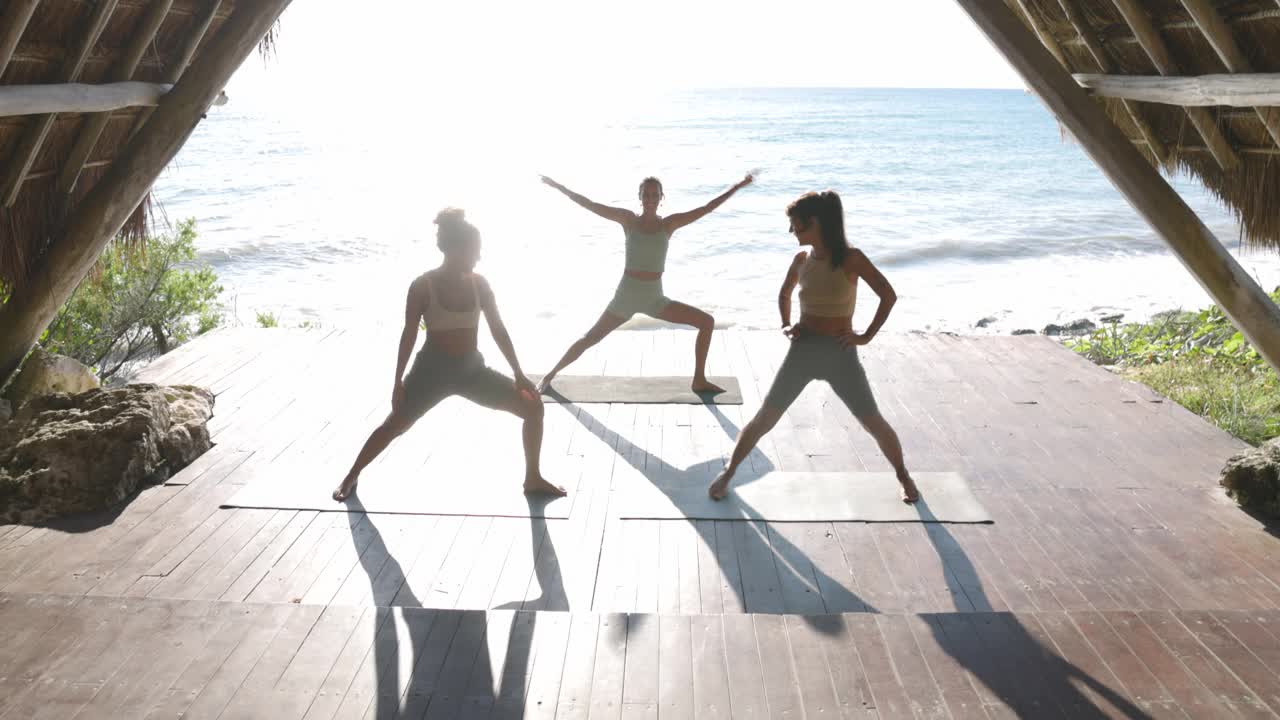 在热带度假胜地的海滨亭子里，在课堂上练习瑜伽的妇女的宽镜头高角度视频素材
