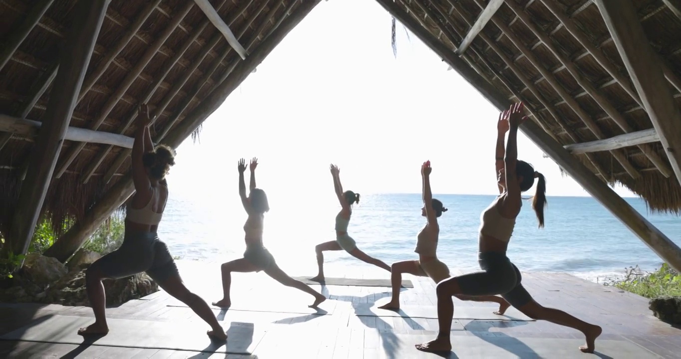 在热带度假胜地的海滨亭子里，在课堂上练习瑜伽时，女人们摆出战士的姿势视频下载