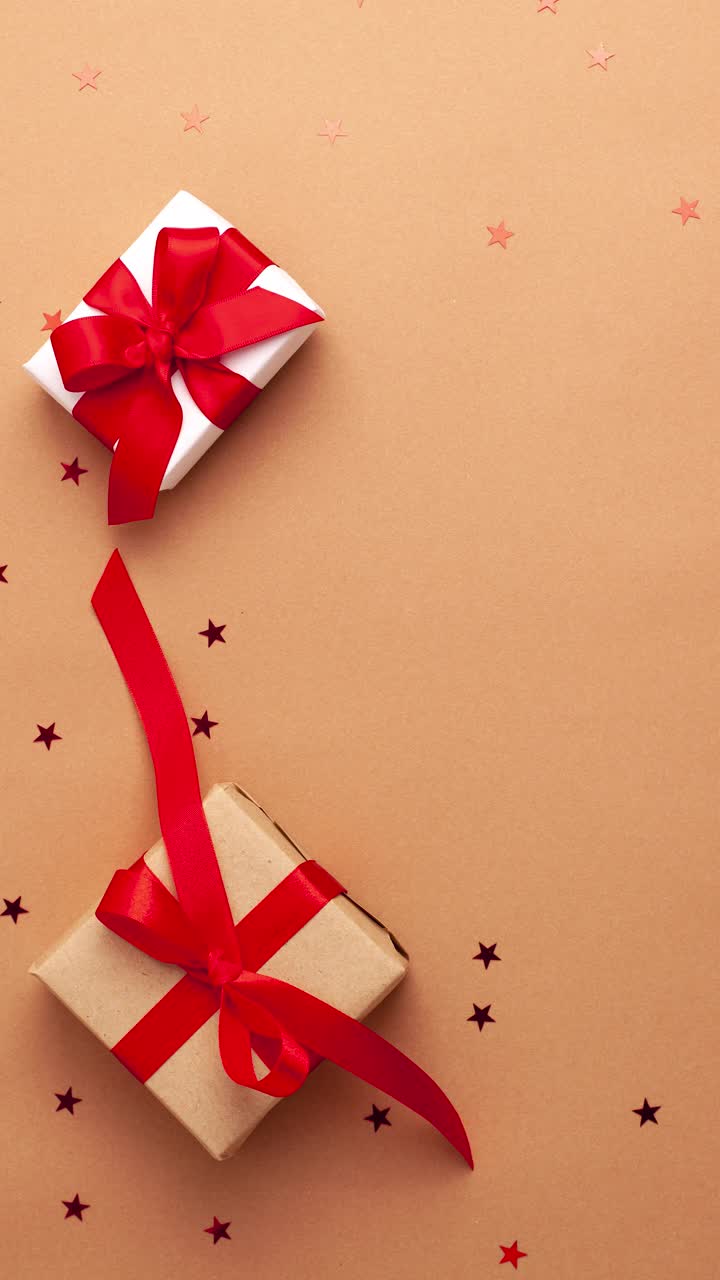 人的双手在白色毛衣带走白色和棕色的纸礼物与红色缎带蝴蝶结在棕色的背景与红色的星星形状。停止运动垂直动画圣诞节假期平lay视频素材