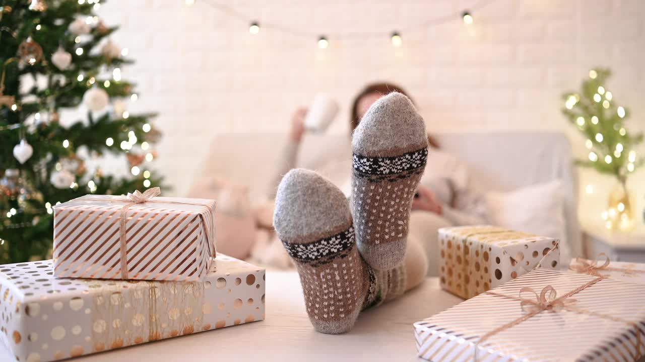 一个女人坐在圣诞树旁的沙发上。女人们把腿、脚放在冬季羊毛袜的桌子上，并在包装完手工礼盒后放松。提出了家庭。视频下载