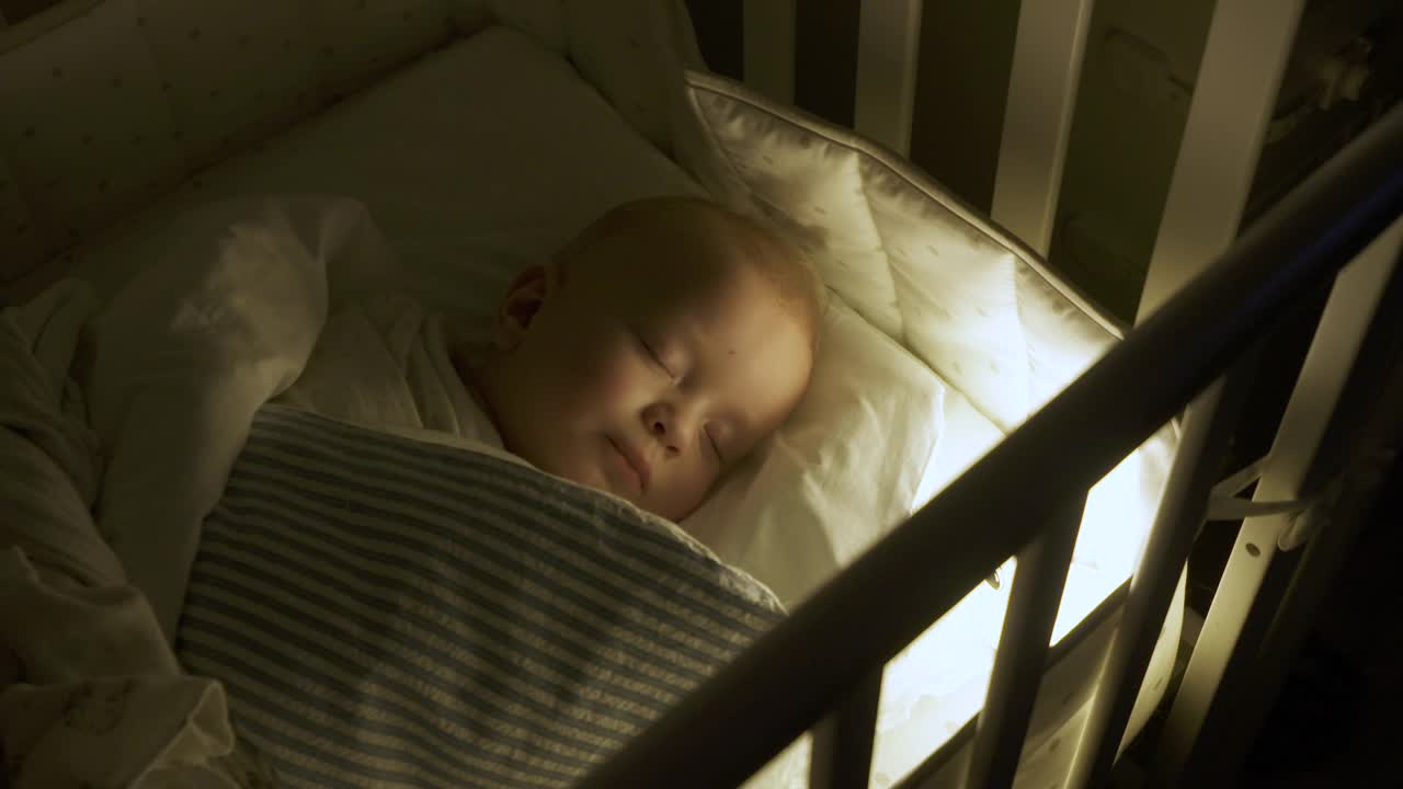 婴儿襁褓躺在婴儿床，9个月的男婴睡在夜灯下。视频下载