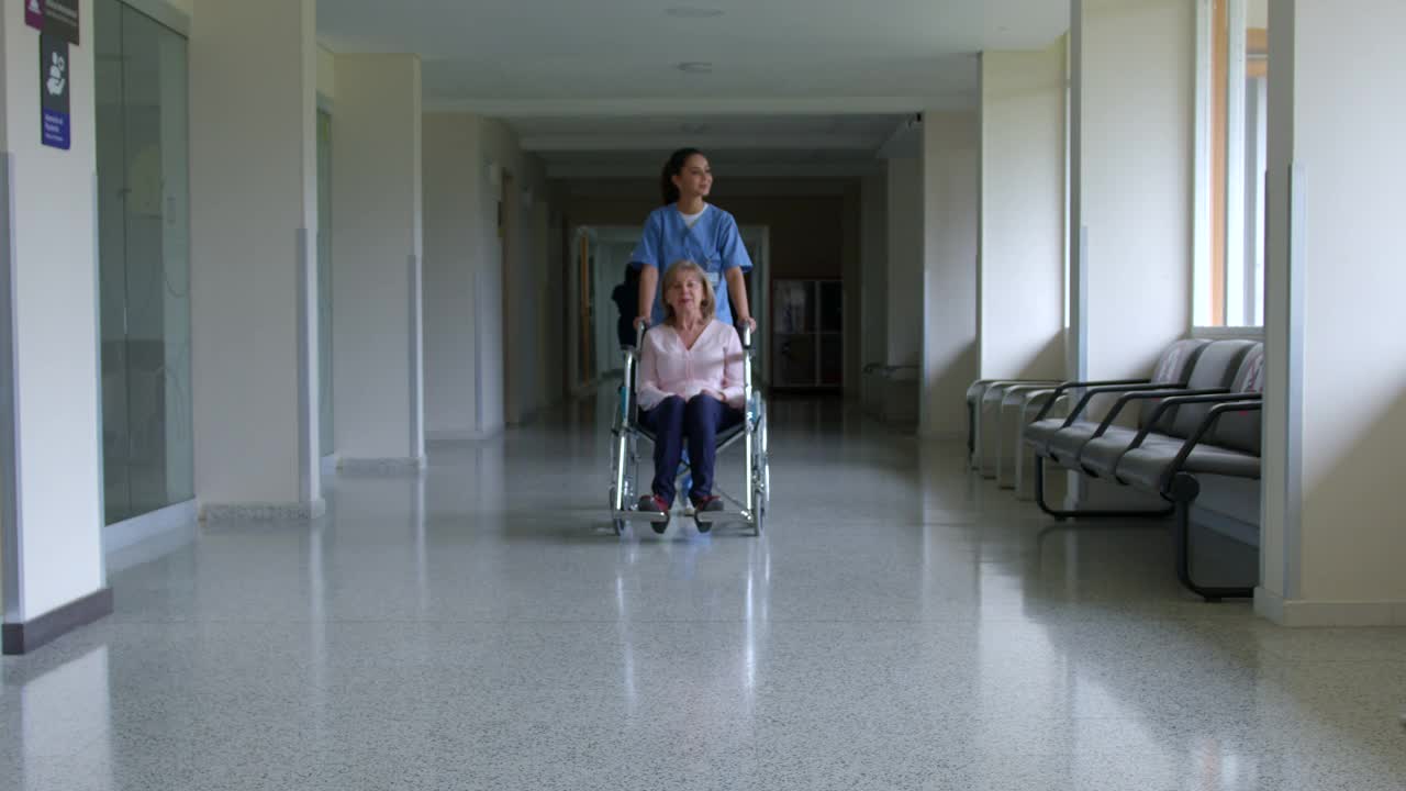 友好的护士走过医院的走廊，带着一个坐轮椅的老年女性病人，一边指着一边给她看东西视频素材