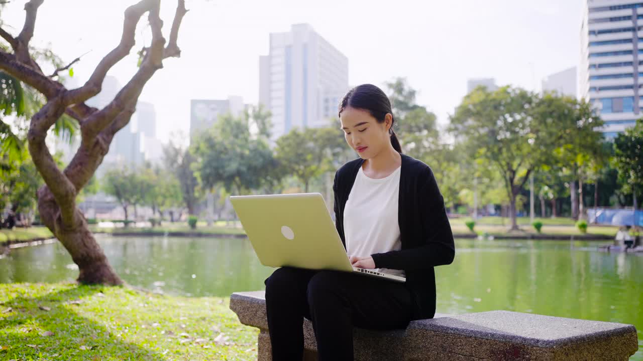 年轻的亚洲女商人拿着一杯咖啡，用笔记本电脑在现代城市的公园里工作视频素材