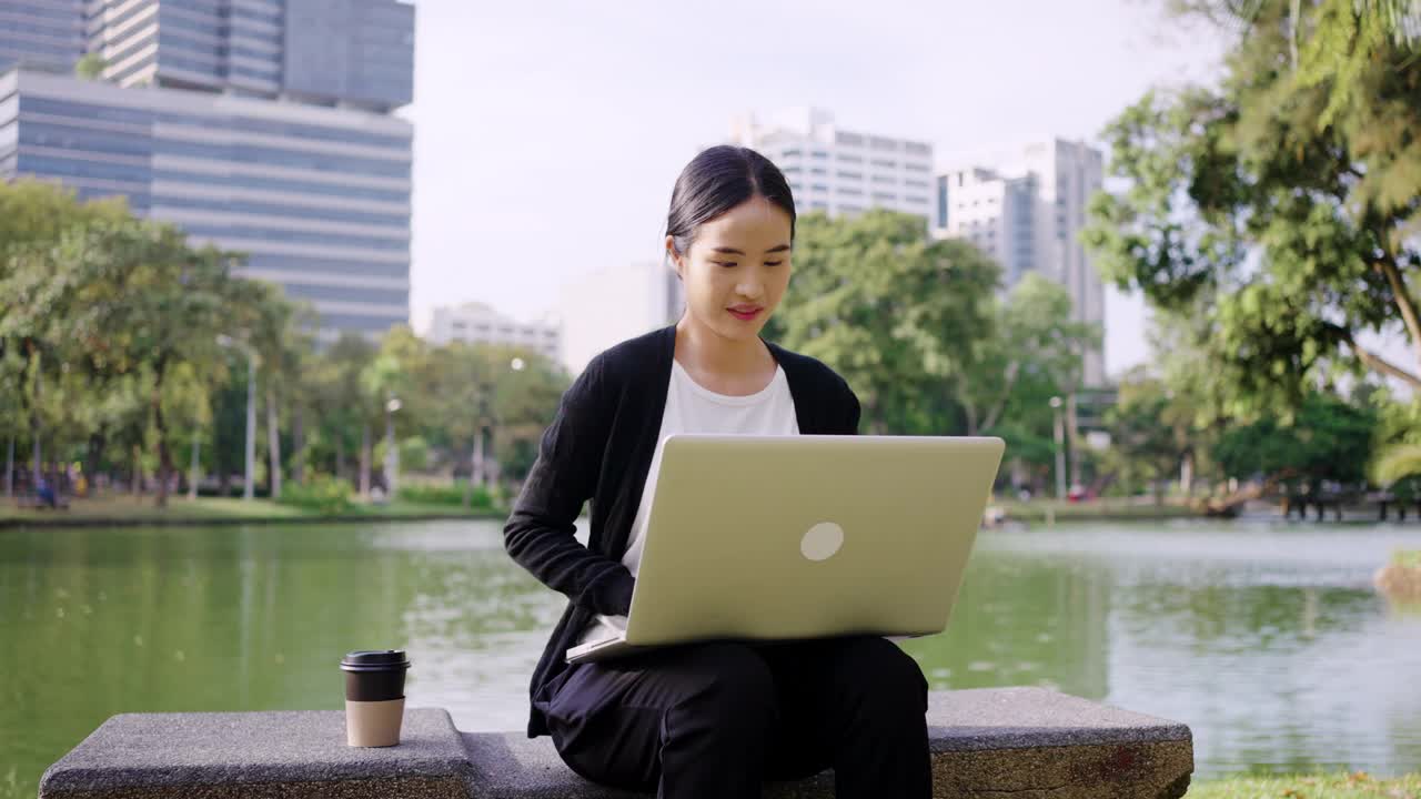 年轻的亚洲女商人拿着一杯咖啡，使用笔记本电脑在现代城市的一个公园户外工作视频素材