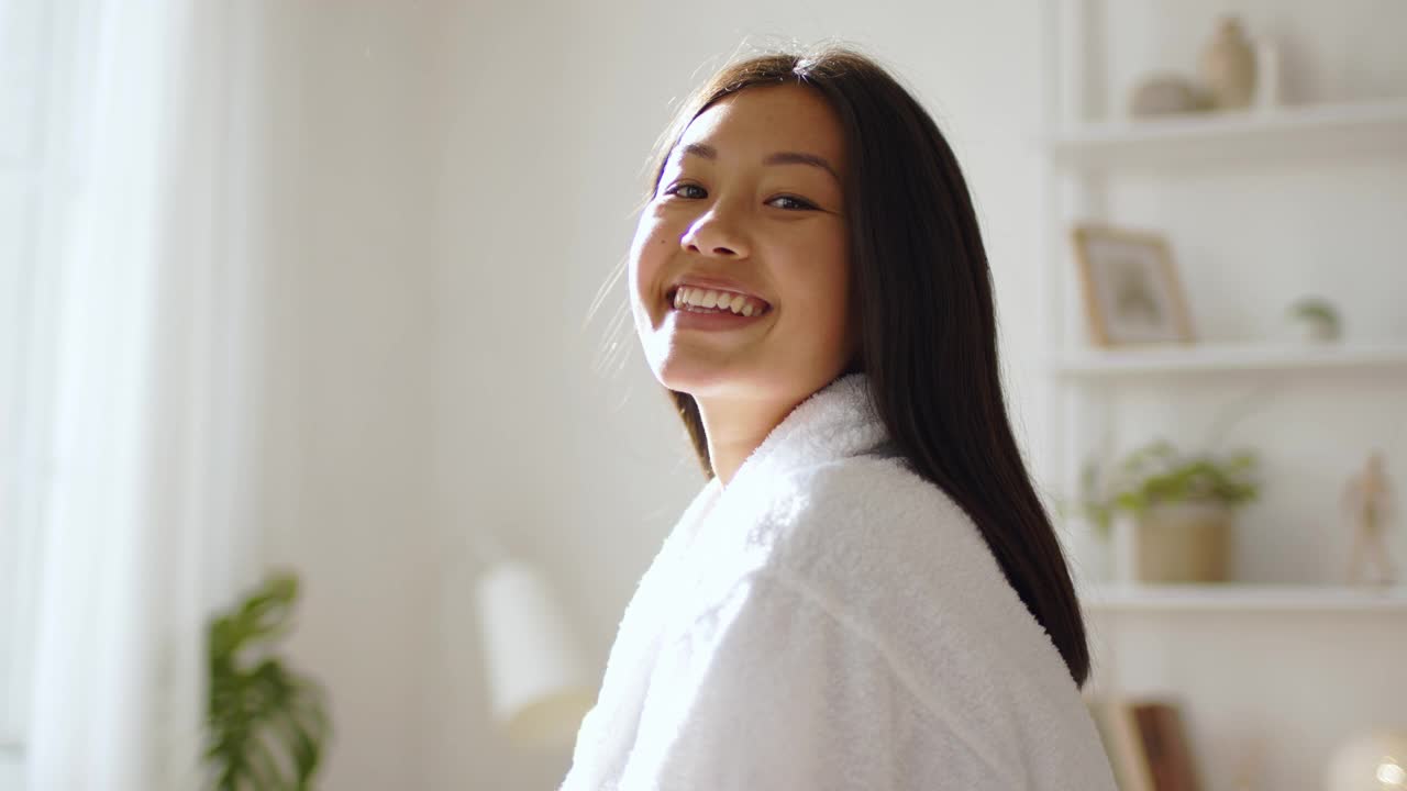 年轻和平的韩国妇女穿着白色的浴衣对着镜头微笑，在早晨的阳光下，在家里摆姿势，慢动作视频素材