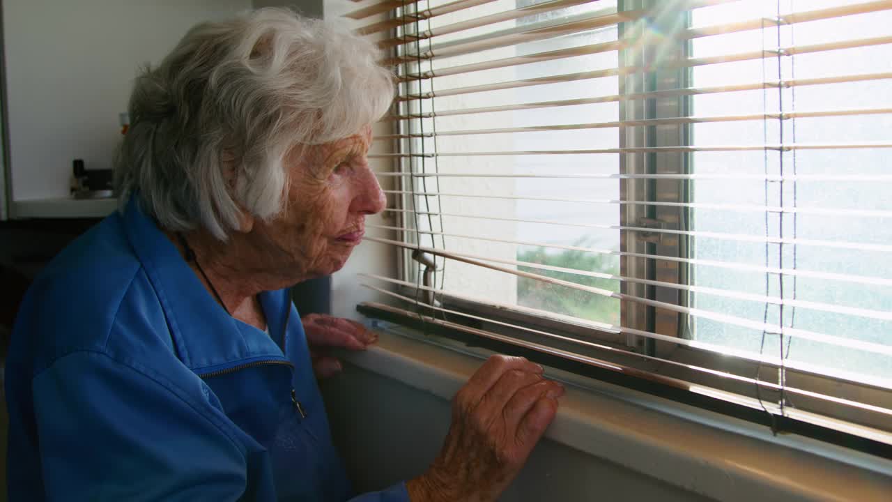 一位年长的白人妇女站在室内，透过百叶窗往窗外看视频下载