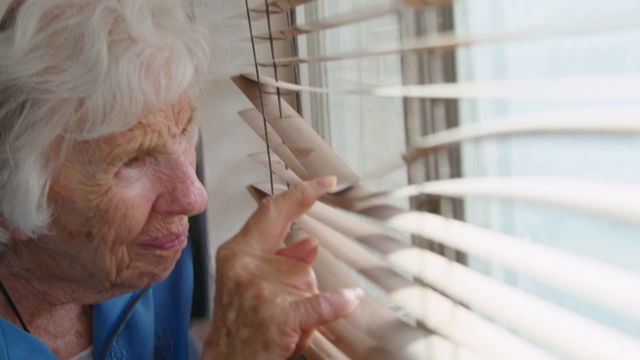 特写镜头:一个忧虑的、孤独的老年白人妇女站在室内，透过百叶窗望向窗外，试图看到外面发生了什么视频下载