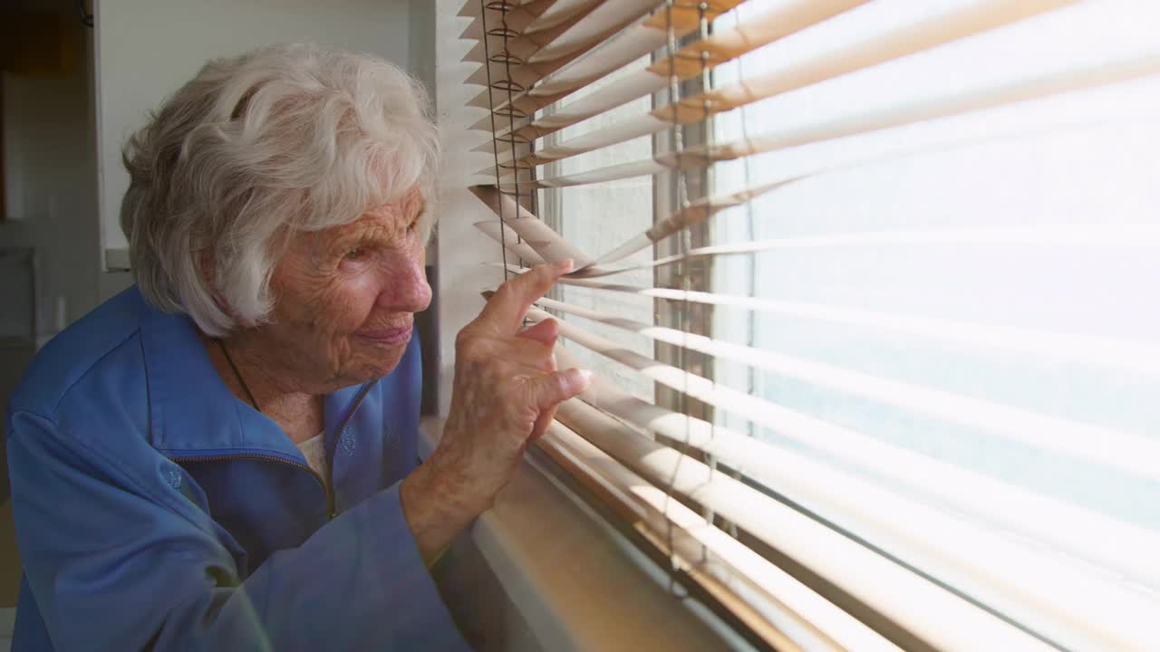 忧虑，孤独的老年白人妇女站在室内透过百叶窗望向窗外视频下载