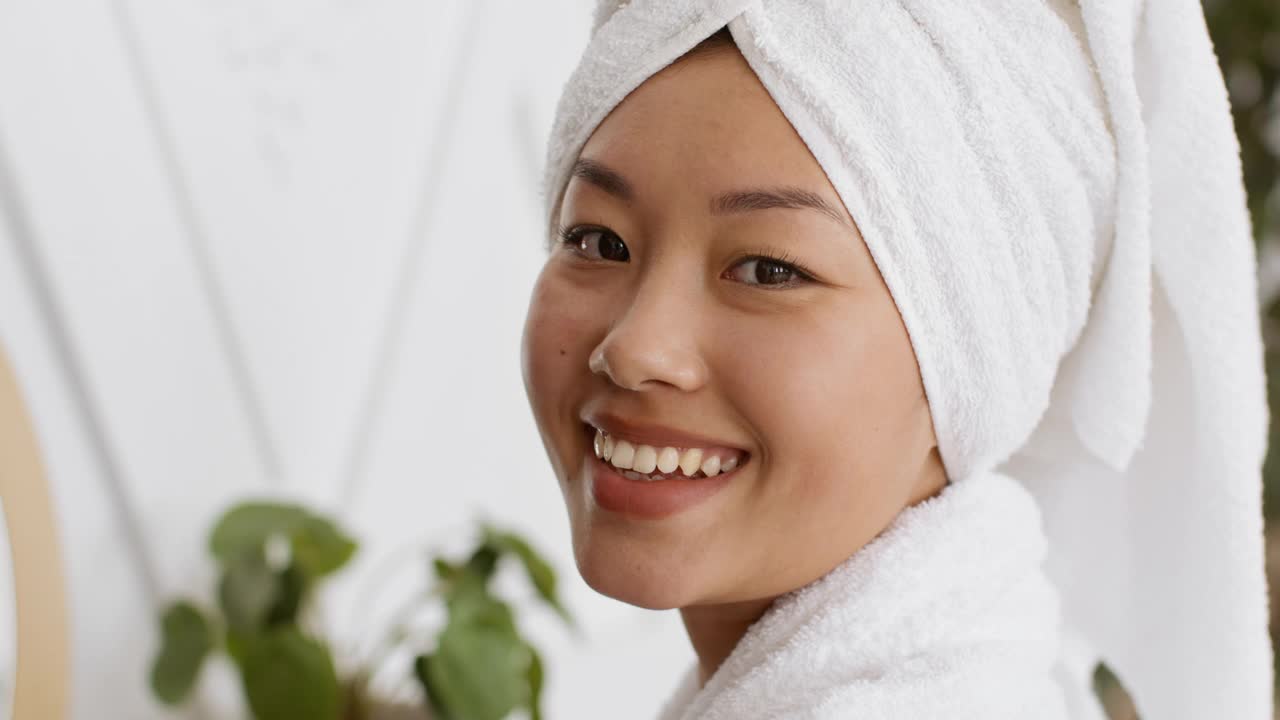 健康的概念。愉快的亚洲妇女头戴毛巾对着镜头微笑，在脸上涂抹保湿霜视频素材