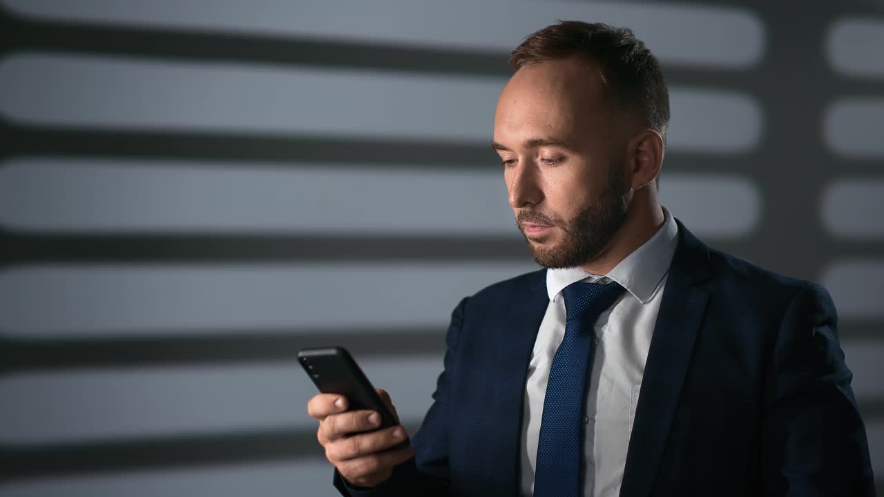 自信的商人聊天打字信息使用智能手机晚上办公室灰色房间百叶窗阴影视频素材