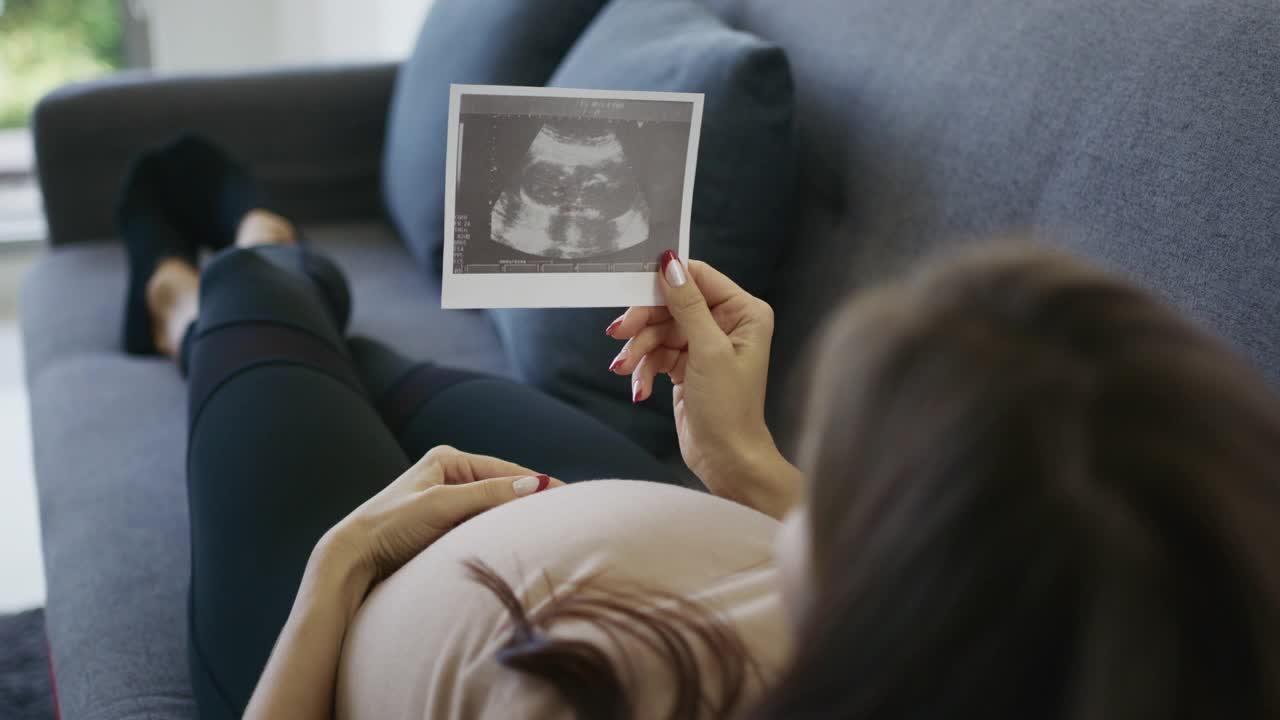 母亲躺在沙发上，看着她怀孕的样子，同时抱着她孩子的超声波图像视频下载