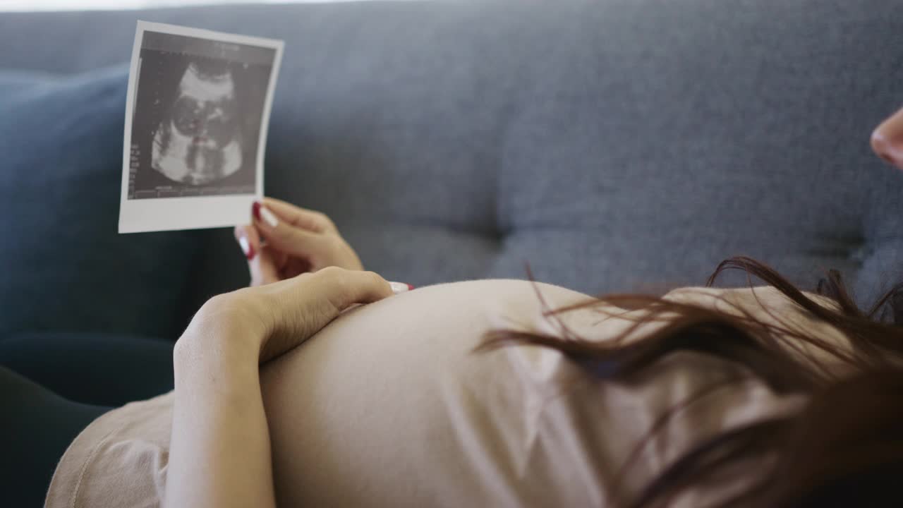 母亲躺在沙发上，看着她怀孕的样子，同时抱着她孩子的超声波图像视频素材