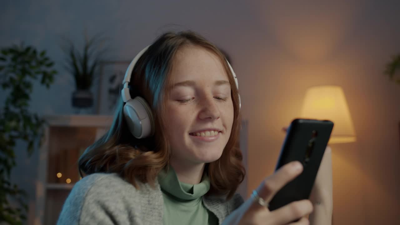 慢动作可爱的女孩在黑暗的房间里戴着耳机听音乐和使用智能手机视频素材