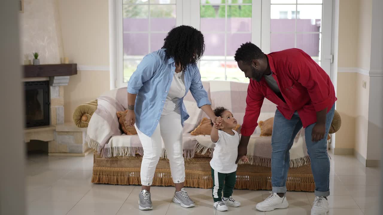 宽镜头快乐的非裔美国父亲，母亲和蹒跚学步的儿子在家里的客厅跳舞。积极自信的千禧一代夫妇和可爱的男孩周末在室内玩。视频素材