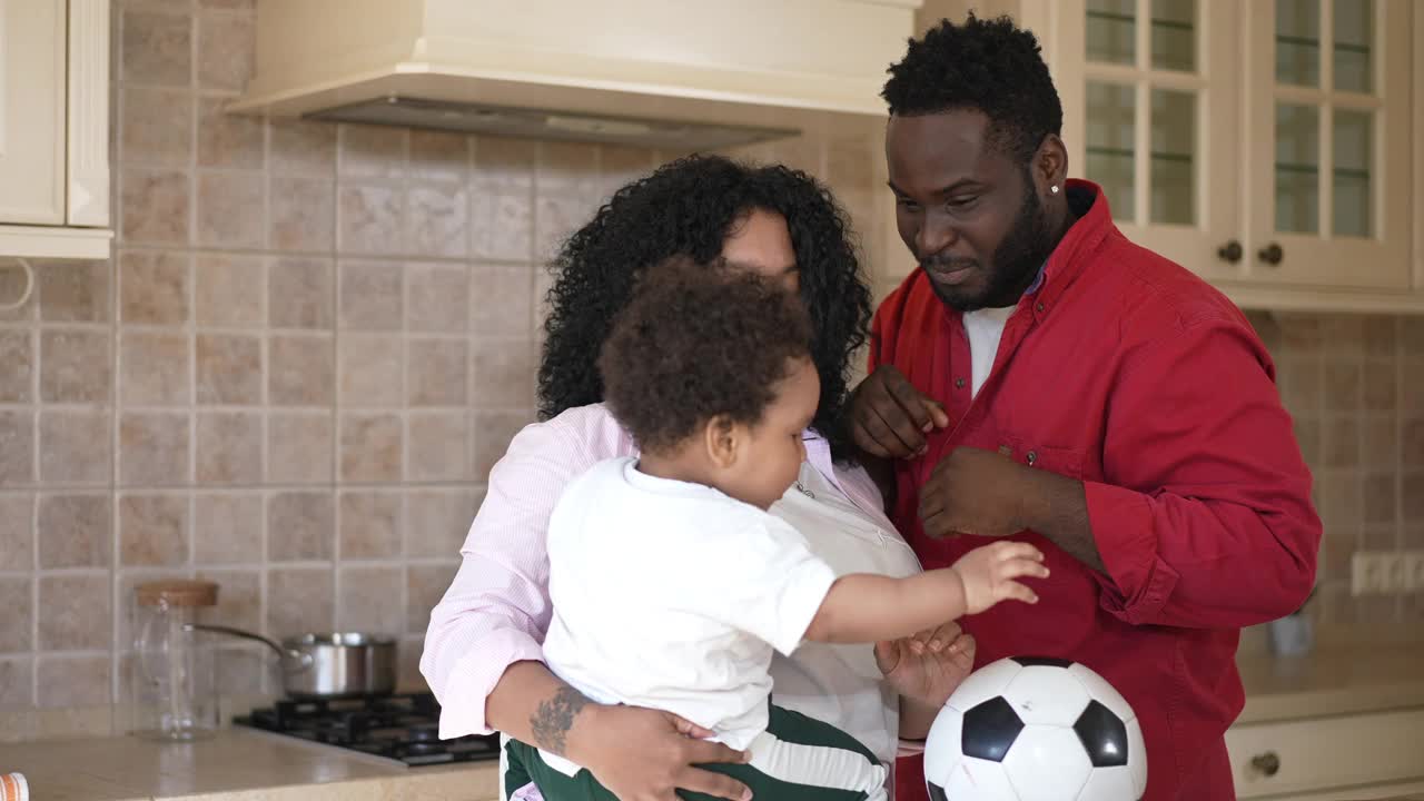 蹒跚学步的男孩用慢动作把足球传给快乐的年轻非洲裔美国父亲，就像快乐的母亲羡慕孩子一样。轻松自信的家庭享受阳光明媚的早晨在厨房在家里。视频素材