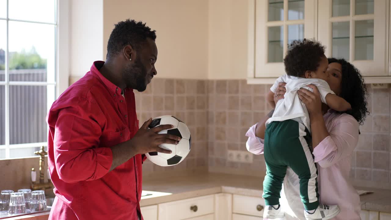 侧面视图肖像正面年轻的非洲裔美国父亲与足球谈话蹒跚学步的儿子在母亲的手搔痒孩子。快乐的帅哥美女和可爱的男孩在厨房在家里。视频素材