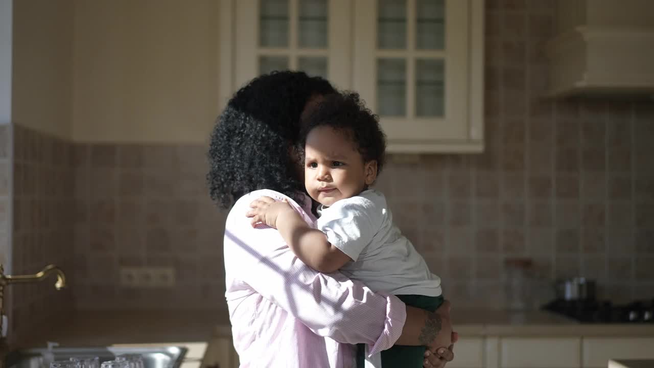 边看快乐的非洲裔美国年轻母亲亲吻儿子作为父亲带来足球。可爱的夫妇快乐的父母享受周末小可爱的男孩在家里的厨房室内。视频素材