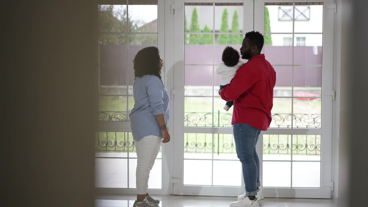 快乐的非裔美国父母欣赏蹒跚学步的儿子在客厅慢动作走。微笑美丽的母亲和英俊的父亲享受周末可爱的孩子在家里。视频素材