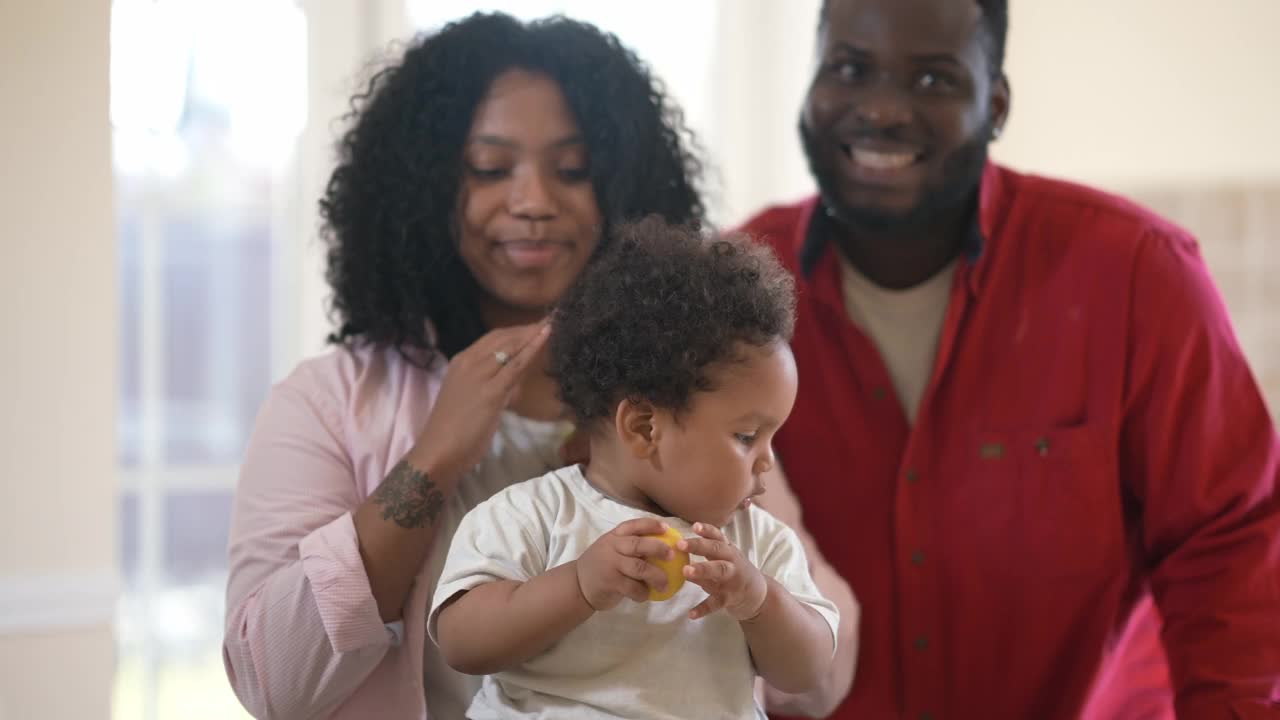 前视图可爱的非洲裔美国男孩玩苹果和柠檬作为模糊夫妇的父母微笑看着相机背景。无忧无虑快乐的蹒跚学步的孩子在家里与父亲和母亲的肖像。视频素材