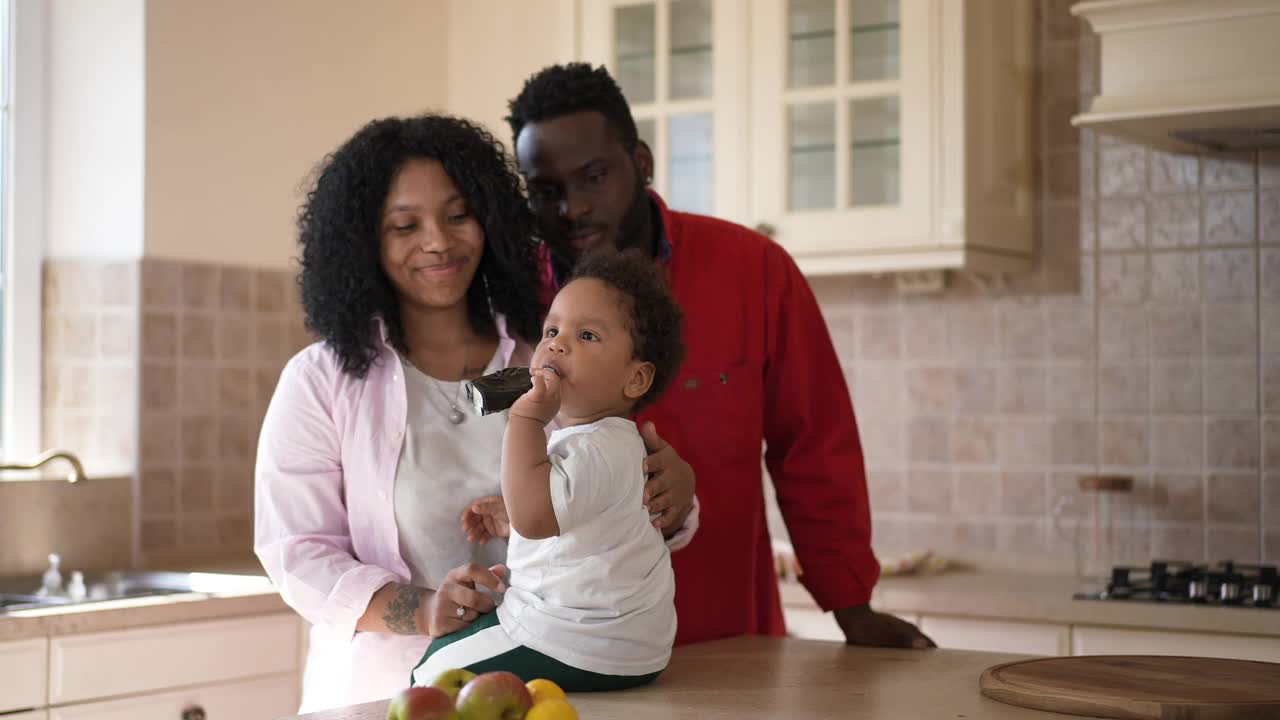 无忧无虑可爱的非洲裔美国幼儿男孩吃儿童食品从管满足父亲亲吻妻子和儿子。轻松愉快的年轻家庭在早上一起休息在家里的厨房。视频素材