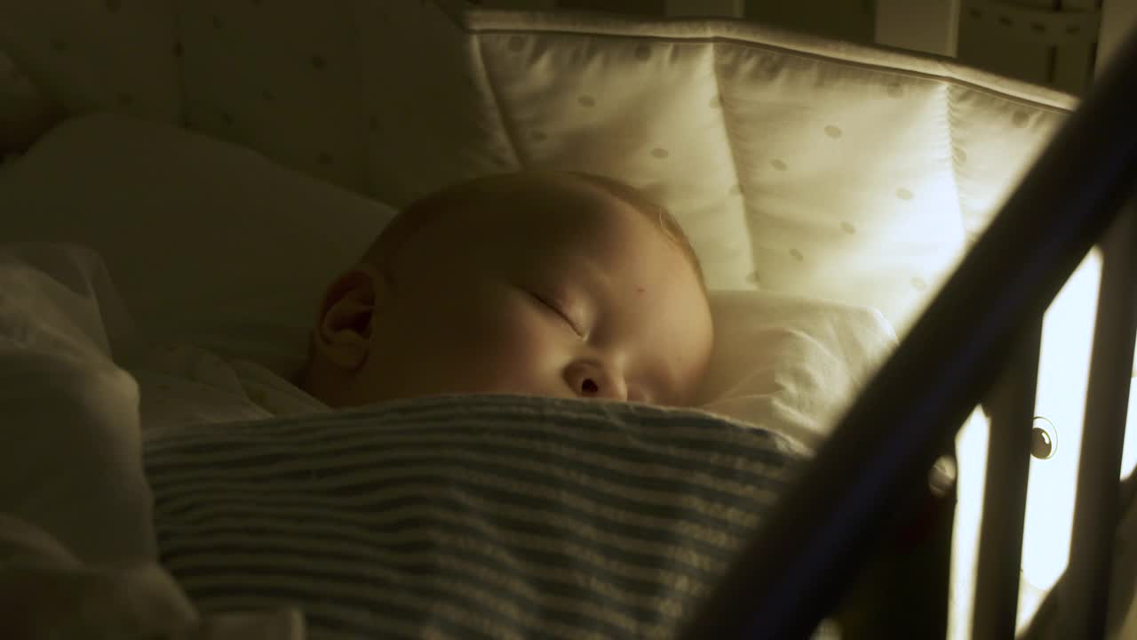 在夜晚的灯光下，襁褓中的9个月男婴睡在婴儿床上。毯子裹着孩子，让他安静下来，让他入睡。视频下载