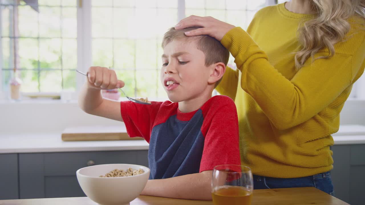 不快乐的儿子在家里吃早餐麦片在厨房柜台，而母亲在给他梳头视频素材