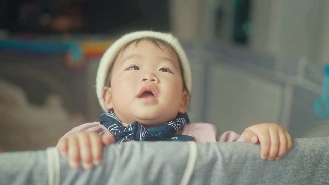 4k亚洲女婴面部表情和积极情绪，穿上冬装，站在客厅家庭生活内的婴儿围栏上视频下载