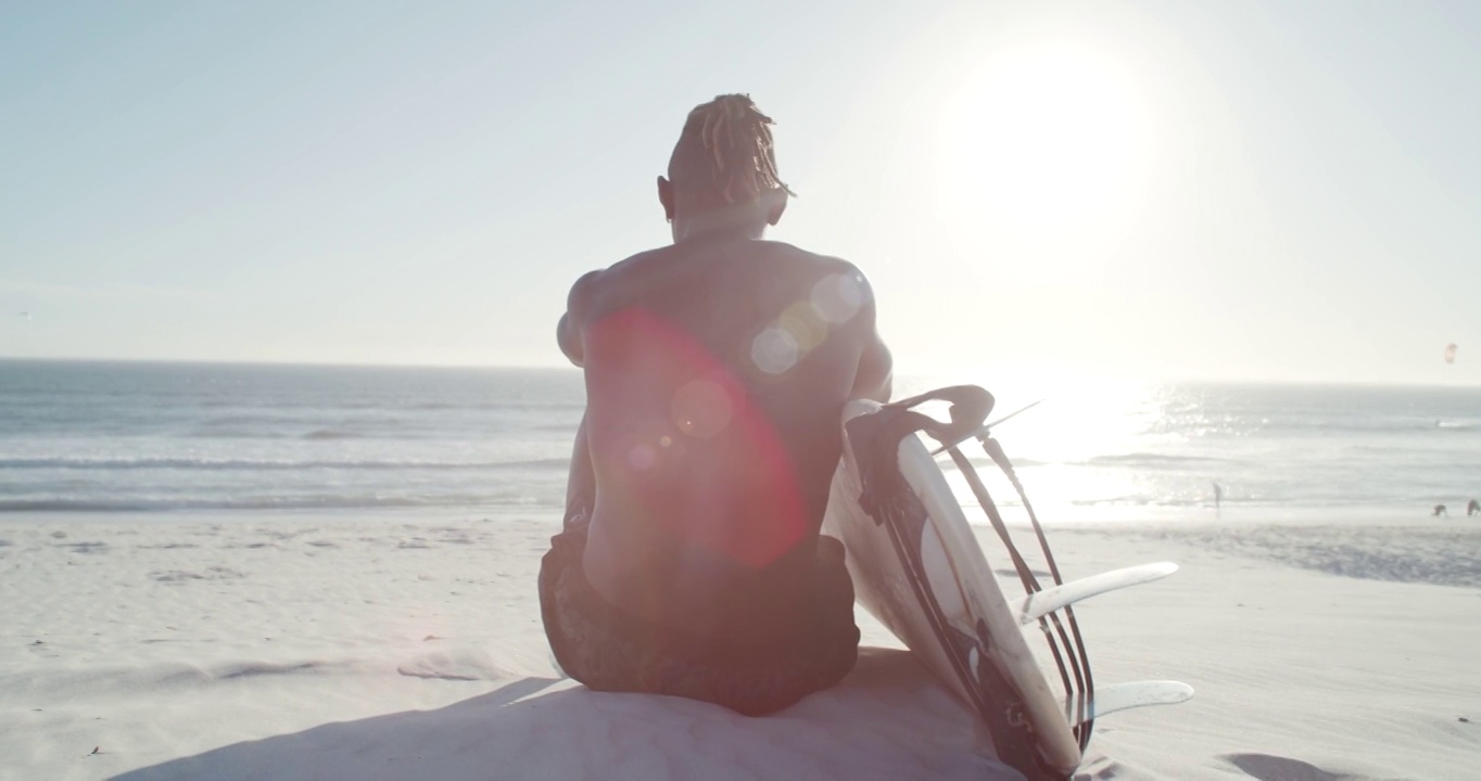一个无法辨认的年轻男性冲浪者带着冲浪板坐在海滩上的4k视频视频下载