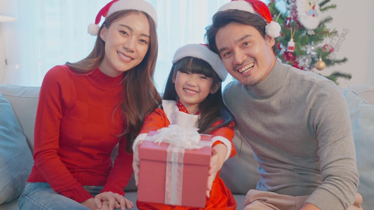 肖像上的亚洲幸福家庭戴着圣诞帽，坐在客厅里。漂亮的父母，父亲，母亲和年幼的女儿在红衣服看相机准备在家里庆祝圣诞节视频素材
