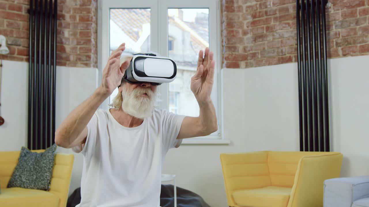 前视图，可爱的活跃的现代70岁的大胡子男人，穿着居家服，戴着增强现实眼镜，在装饰精美的客厅里，在虚拟屏幕上工作视频下载