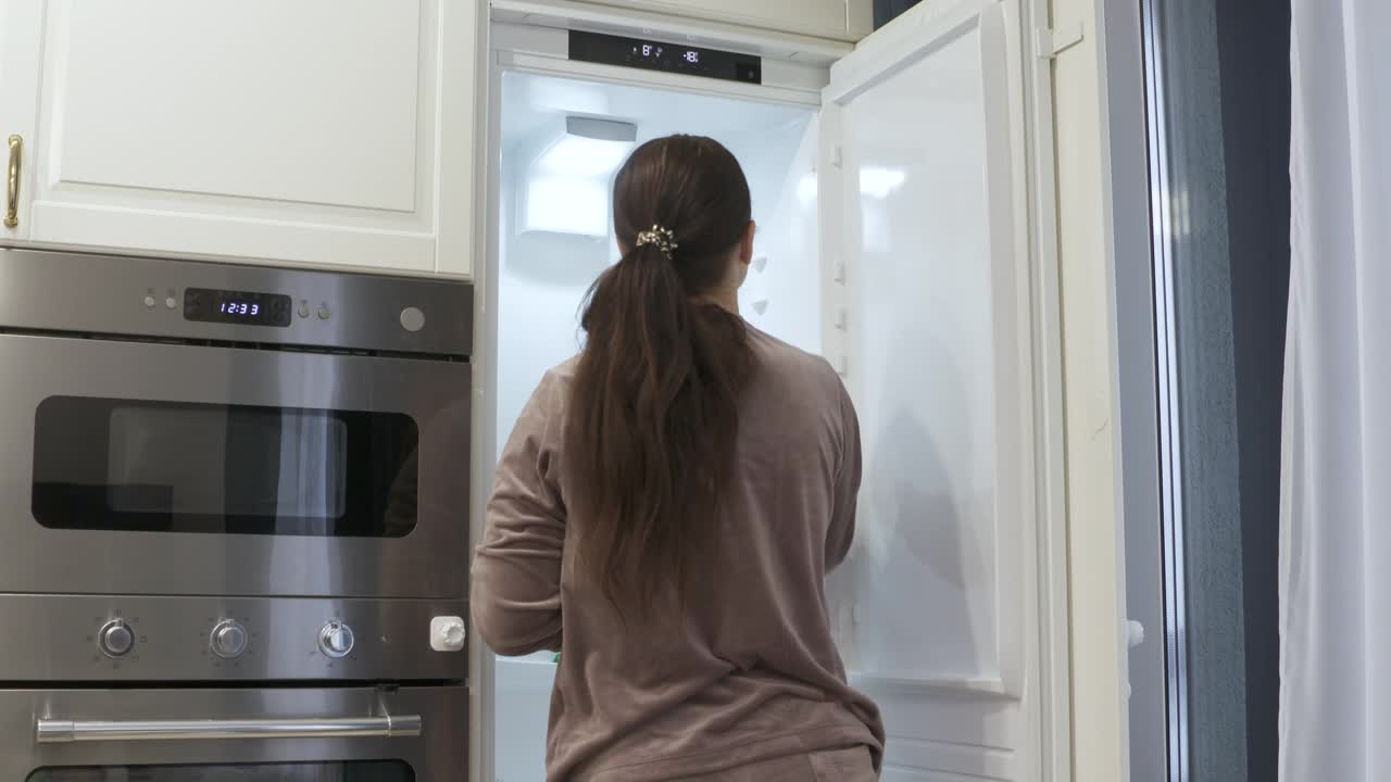 一位妇女正在打扫家里厨房里的冰箱。集成冰箱内建在白色厨房住宅，现代公寓内部。视频素材