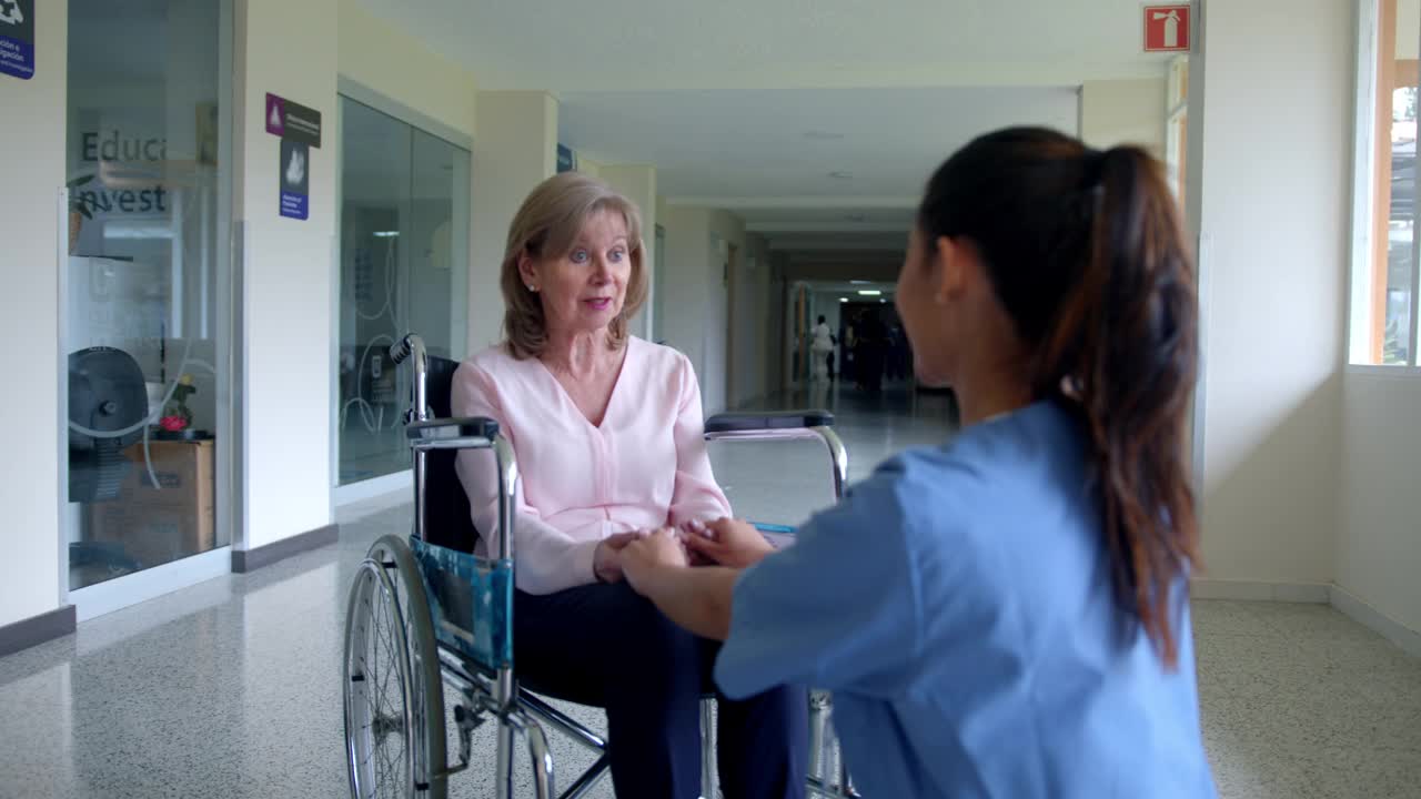 在医院走廊的轮椅上，一位细心可爱的护士握着一位老年病人的手，谈笑风生视频素材