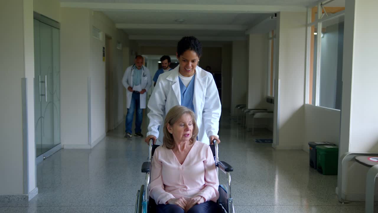 友好的黑人女医生穿过医院的走廊，带着一个坐着轮椅的老年女病人边说边笑视频素材