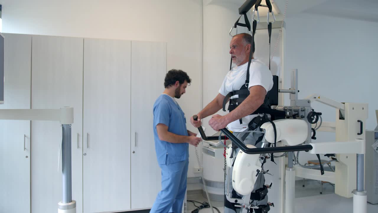 男性治疗师调整跑步机的速度，而老年男性患者是在一个物理恢复中心的动力外骨骼视频素材