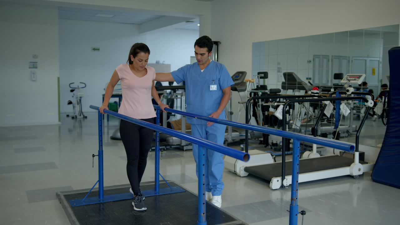 年轻的女性患者在进行物理治疗时，靠双杠努力行走，而男性治疗师走在她旁边视频下载
