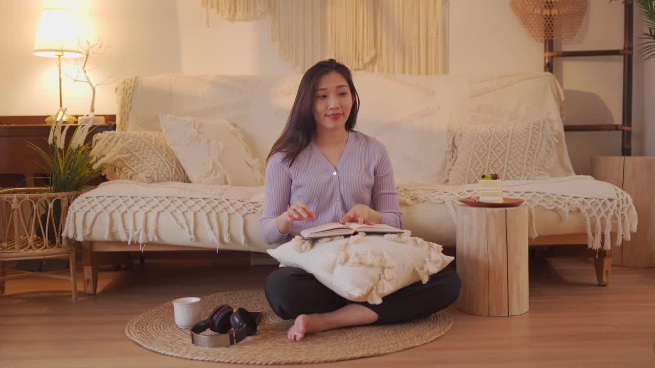 亚洲华人妇女享受她的空闲时间坐在客厅的地面上看书，听音乐，吃蛋糕在周末深夜视频素材