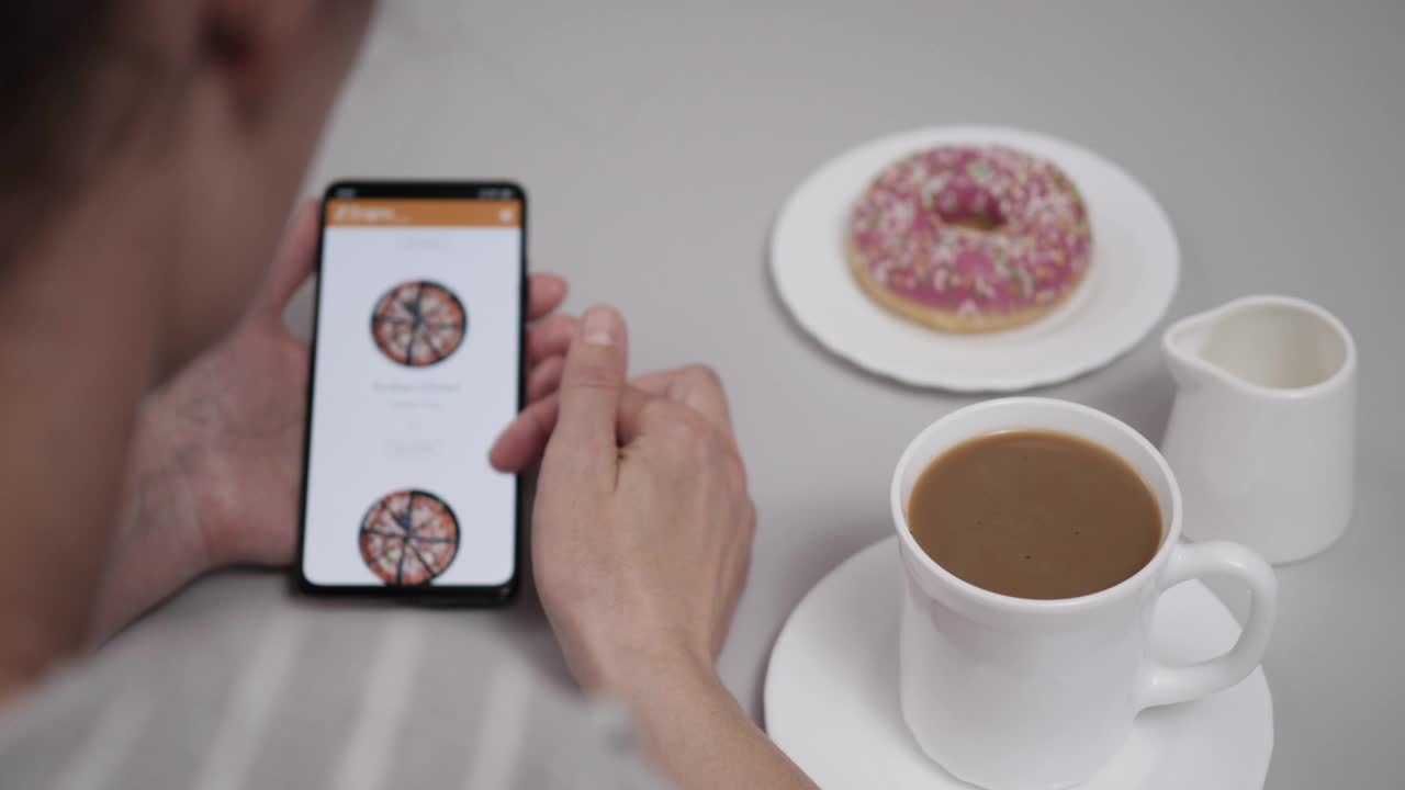 女性用外卖App点披萨。一名女子在网上商店用智能手机订购食物。一个女人坐在餐桌旁，手里拿着咖啡甜甜圈和智能手机。视频下载