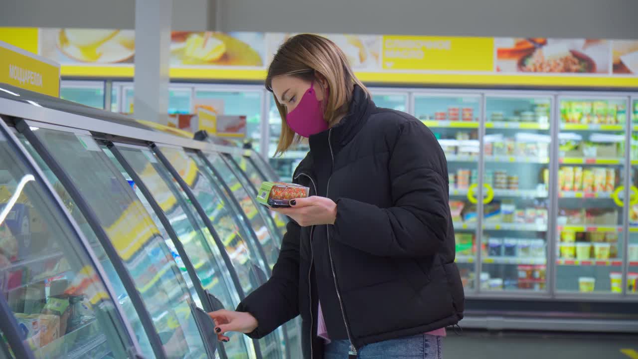 一位戴着口罩保护Covid-19的妇女站在冷藏库的货架前。有选择性的重点视频素材