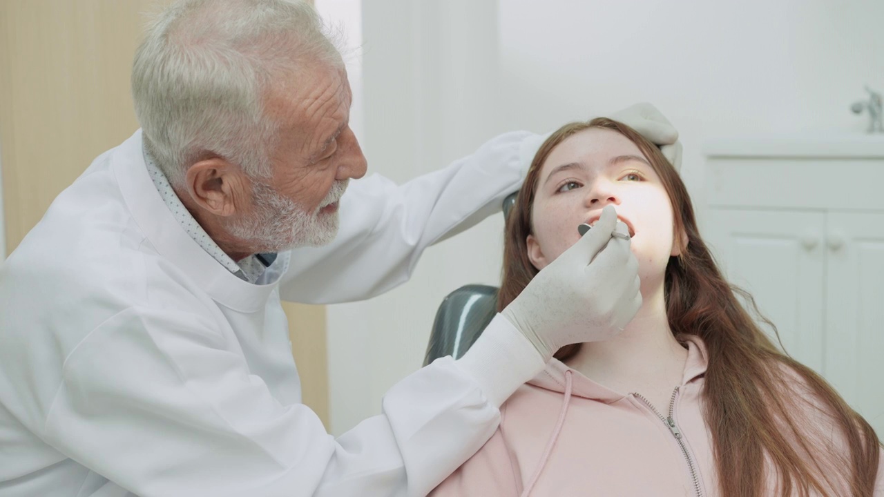 老牙医用牙科设备为十几岁的女孩病人检查和检查牙齿，女孩坐在牙科椅上等待治疗视频素材