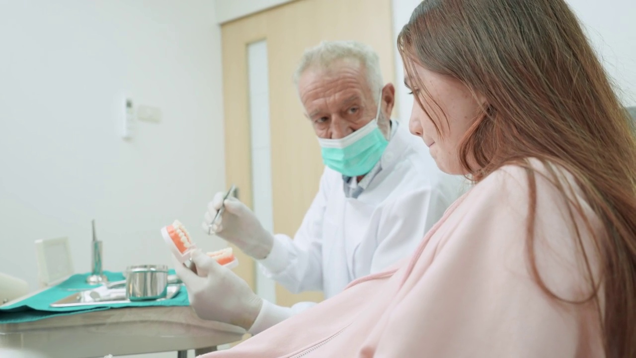 戴着防护口罩的老牙医用口腔和牙齿模型向坐在牙科椅上，向后看的少女病人展示如何正确刷牙视频素材