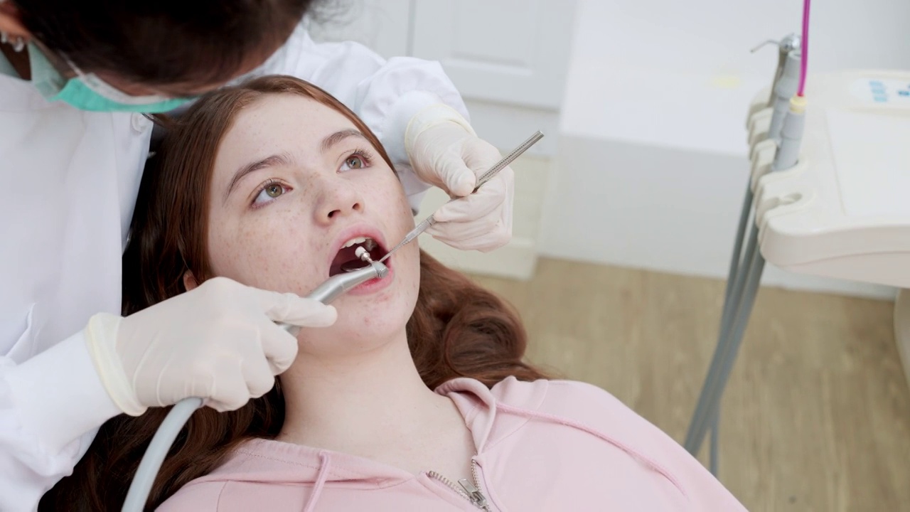 特写:十几岁的小女孩坐在牙科病房，张开嘴，让亚洲中年牙医用斜镜、吸唾液等牙科设备检查和补牙视频素材