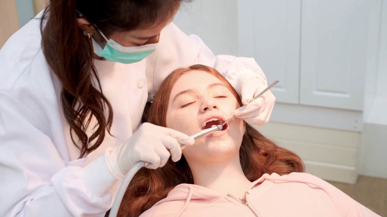 亚洲中年女牙医用斜面镜、吸唾液器等牙科设备为少女病人做牙科检查和牙齿清洁，少女坐在带照明的牙科椅上视频素材