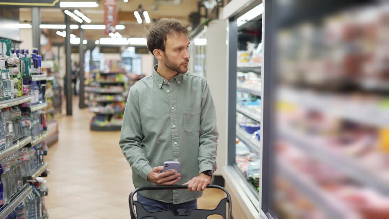穿着衬衫的快乐男人推着手推车穿过超市挑选食品。检查手机上的购物清单视频素材