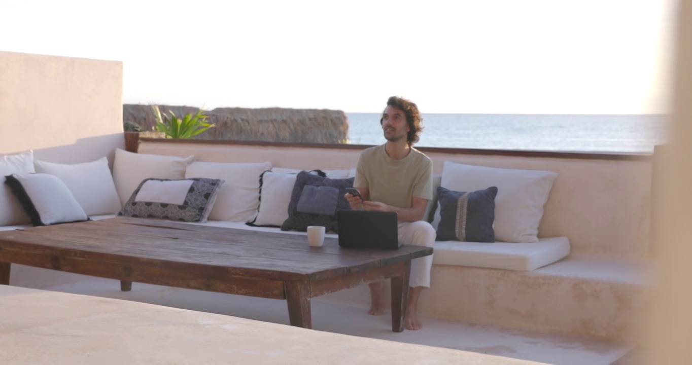 在日出时，微笑的男人在豪华的热带海滩别墅甲板上检查手机和笔记本电脑工作的宽镜头视频下载