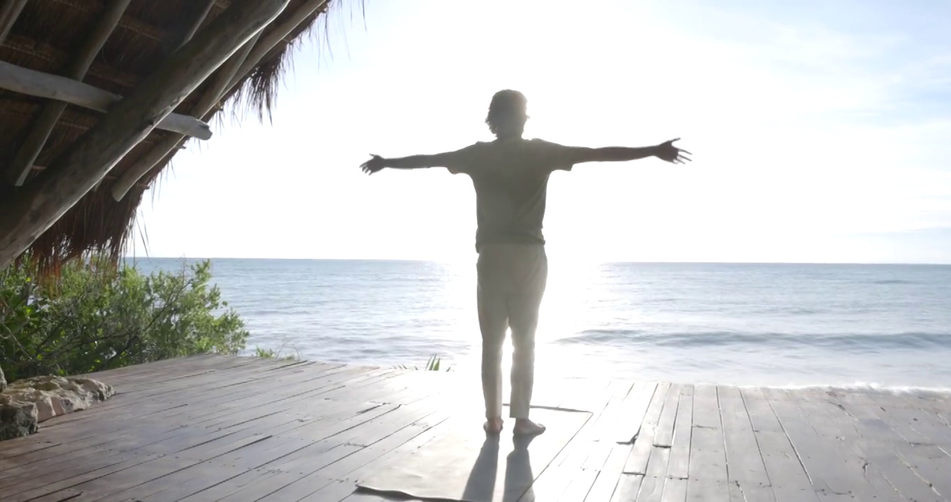 日出时分，在豪华的热带度假胜地的海滨亭子里，男人在练习瑜伽前伸展的宽镜头低角度视频下载