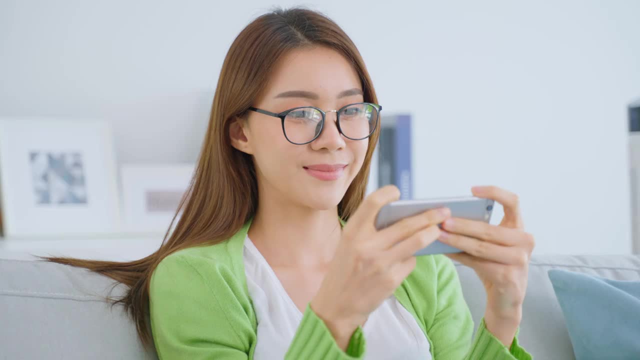 亚洲美女戴眼镜，在家用手机聊天。有吸引力的年轻女孩坐在沙发上，感觉兴奋和快乐后使用智能手机检查在客厅的好消息。视频素材
